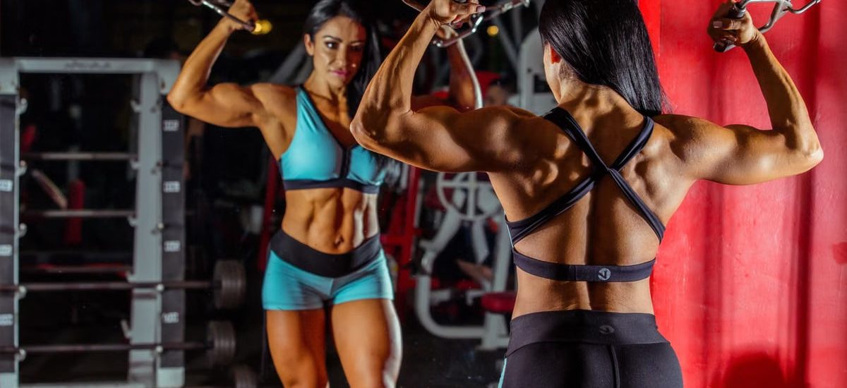 Strong female bodybuilder doing cable workout in a gym with mirror reflection.